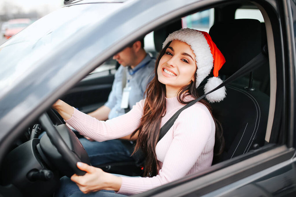 Picture of attractive young woman sit on drivers place. Hold hands on steering wheel and smile. Christmas or new year time. Wear red hat and smile. Man sit beside. - Fotografie, Obrázek