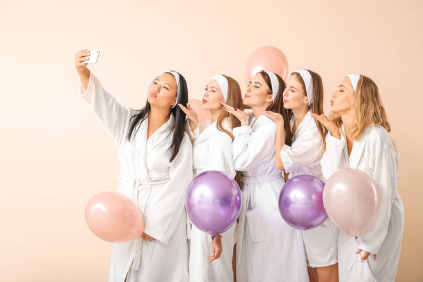 Beautiful young women in bathrobes and with air balloons taking selfie on light background - Photo, Image