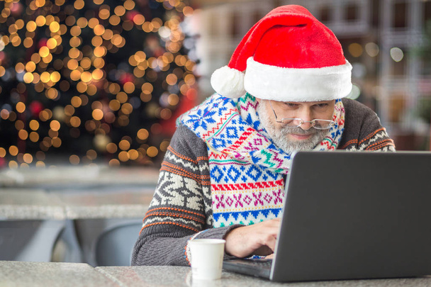 Modern Santa Claus using working on laptop with christmas tree on the background with lights froming 2020 year concept - Foto, afbeelding