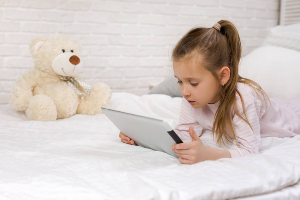 χαριτωμένο μικρό κορίτσι κείτεται στο κρεβάτι χρησιμοποιεί ψηφιακό δισκίο. - Φωτογραφία, εικόνα