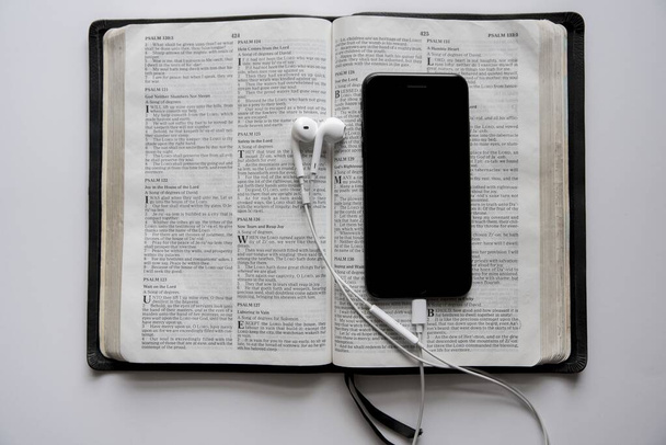 Nahoře záběr smartphonu a sluchátek na otevřené bibli na bílém povrchu - Fotografie, Obrázek