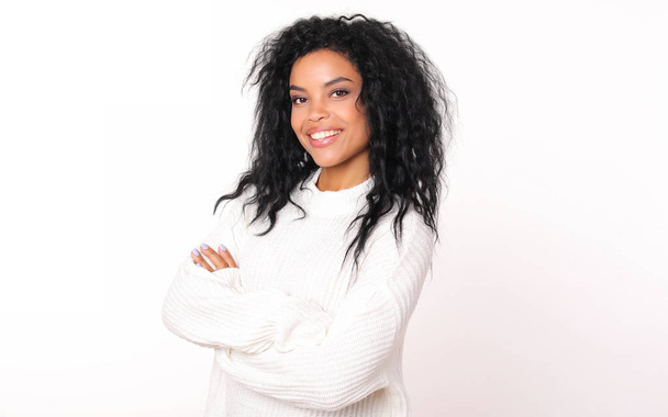 beautiful mixed race woman wearing white winter sweater smiling and looking at camera while posing with crossed arms on white studio background  - Foto, imagen