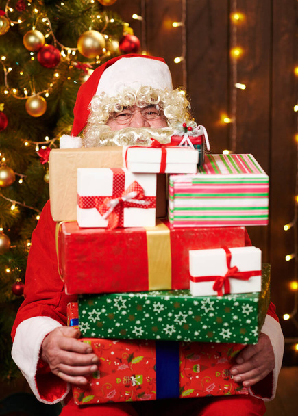 Санта Клаус позує з багатьма подарунками, сидячи в приміщенні біля прикрашеного ксами дерева з сяйвом - Веселого Різдва і Веселих свят.! - Фото, зображення