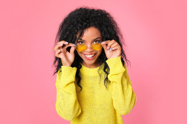 beautiful mixed race woman in yellow sweater wearing sunglasses and looking at camera on pink studio background  - Valokuva, kuva