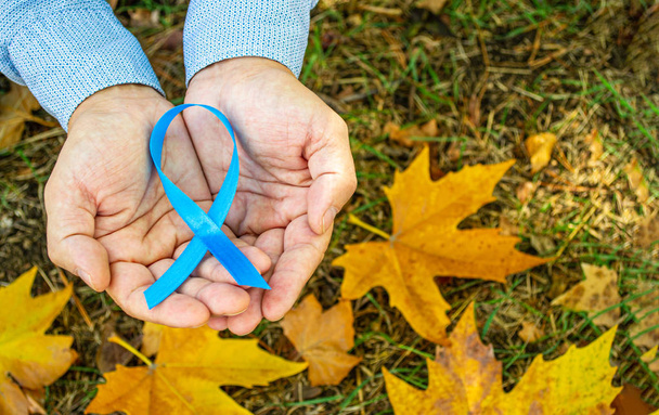  Men's health and prostate cancer awareness in November. Man hands holding a light blue ribbon awareness on the background of autumn maple leaves. A symbol to support men who live with cancer.  Copy space. - Φωτογραφία, εικόνα