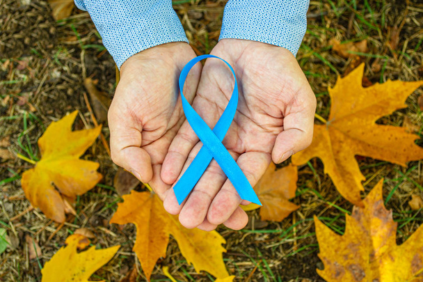  Men's health and prostate cancer awareness in November. Man hands holding a light blue ribbon awareness on the background of autumn maple leaves. Men's health and the concept of world cancer day. - Φωτογραφία, εικόνα
