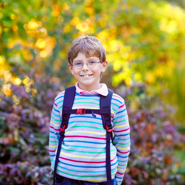 Gelukkig kind jongetje met bril en rugzak of tas op zijn eerste dag naar school op zonnige herfstdag. Kind buiten met gele en lees maple bomen op de achtergrond, terug naar school concept - Foto, afbeelding