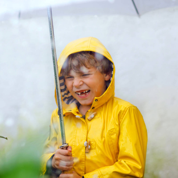Όμορφο μικρό αγόρι στο δρόμο για το σχολείο με τα πόδια κατά τη διάρκεια sleet, δυνατή βροχή και χιόνι με μια ομπρέλα την κρύα μέρα. Ευτυχισμένο και χαρούμενο παιδί με πολύχρωμα κίτρινα ρούχα μόδας παλτό casual - Φωτογραφία, εικόνα
