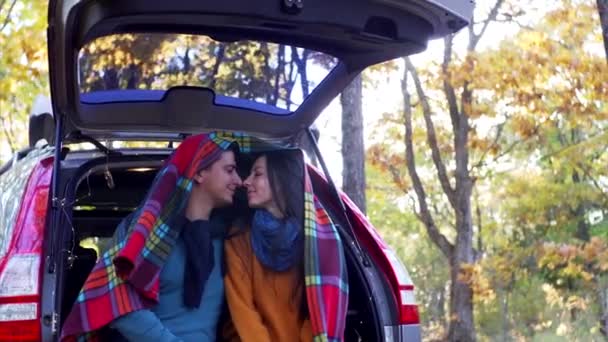 Νεαρός άνδρας και γυναίκα απολαμβάνουν ο ένας τον άλλον στο πορτ-μπαγκάζ του αυτοκινήτου κάτω από ζεστό καρό. Ηλιόλουστο φθινόπωρο - Πλάνα, βίντεο