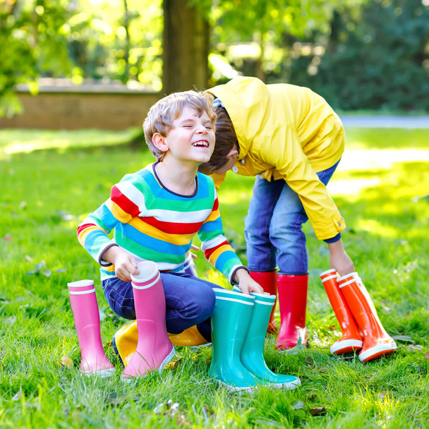 Δύο μικρά παιδιά αγόρια, χαριτωμένα αδέλφια με πολλές χρωματιστές μπότες βροχής. Παιδιά με διαφορετικές μπότες και μπουφάν. Υποδήματα για βροχερή πτώση. Υγιή δίδυμα και οι καλύτεροι φίλοι διασκεδάζουν σε εξωτερικούς χώρους - Φωτογραφία, εικόνα