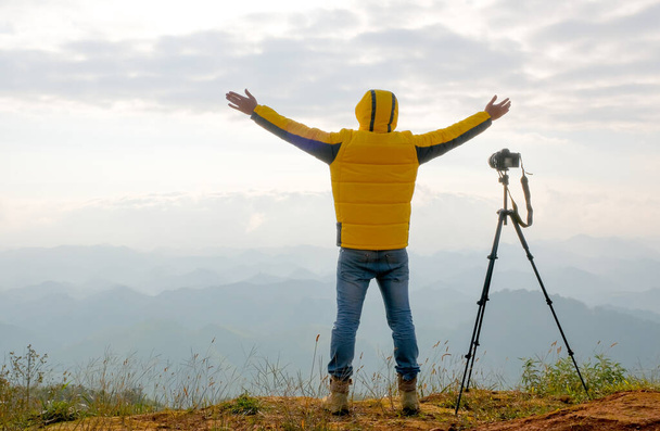Fotograf w żółtej kurtce stoi blisko aparatu i stoi w przednim klifie, zachmurzony i pokazać działania szczęśliwy. - Zdjęcie, obraz