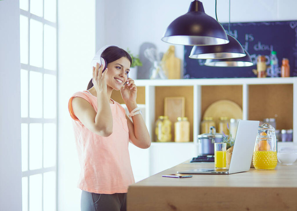 Portrait of a cheerful young woman listening to music with headphones and using laptop computer while standing at the kitchen - Foto, imagen