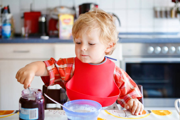 Roztomilý batole chlapec jíst zdravé porrige z lžíce na snídani s jahodovým džemem. Roztomilé šťastné dítě v barevné pyžamo sedí v kuchyni a učí se pomocí lžíce. - Fotografie, Obrázek