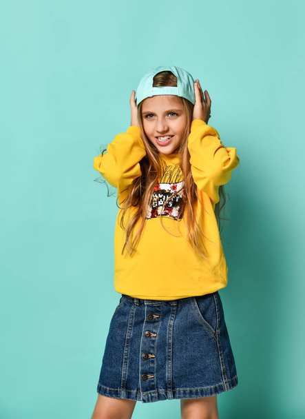 Funny teenage girl in a baseball cap and yellow hoodie covered her ears with her hands on a blue background. - Fotografie, Obrázek