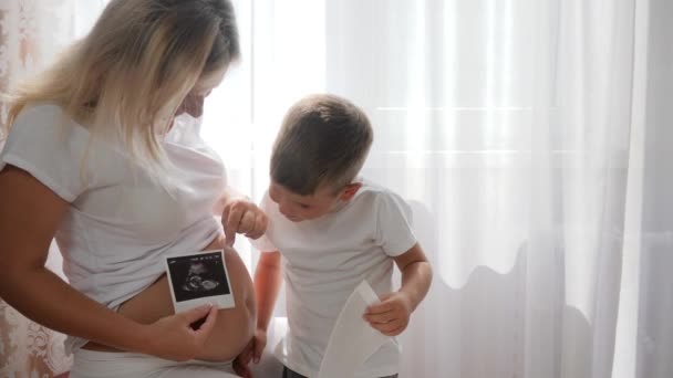 νεαρή έγκυος μητέρα με γλυκό παιδί βλέποντας φωτογραφία υπερηχογραφία στο σπίτι σε έντονο φυσικό φως - Πλάνα, βίντεο
