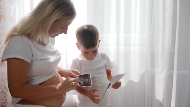mladá těhotná matka se sladkým dítětem sledování fotografie sonografie doma v jasném přirozeném světle - Záběry, video