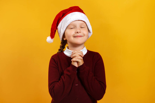 Cute little girl in santa hat is waiting for christmas. The child closed his eyes and made a wish. Closeup portrait on a yellow background - Fotó, kép