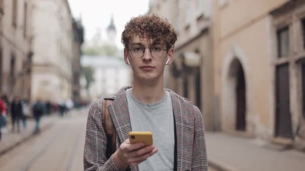 Portrait of Young Guy in Glasses and Earphones Wearing Jacket, Holding Smartphone and Looking to Screen than to Camera at Old City Background. - Video, Çekim