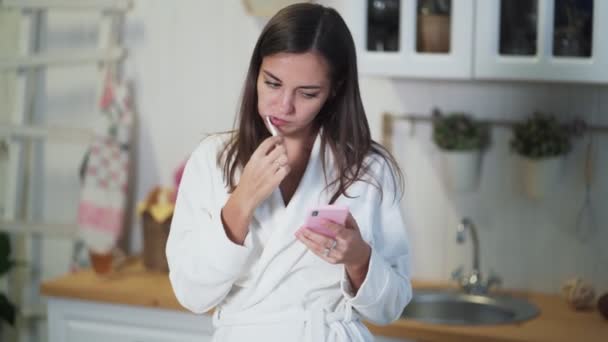 Young woman in white bathrobe brushes her teeth and use smartphone in morning - Video