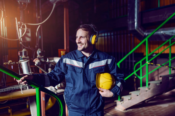 Sonriente trabajador caucásico guapo en traje protector, con antífonas y sombrero de fuerza debajo de la axila de pie en la fábrica
. - Foto, Imagen