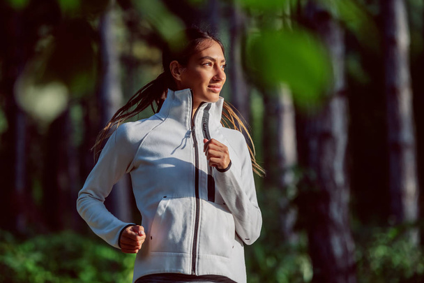 Aantrekkelijke Kaukasische fit sportieve brunette in sweatshirt en met paardenstaart rennend in het bos op zonnige ochtend. Fitness in de natuur. - Foto, afbeelding