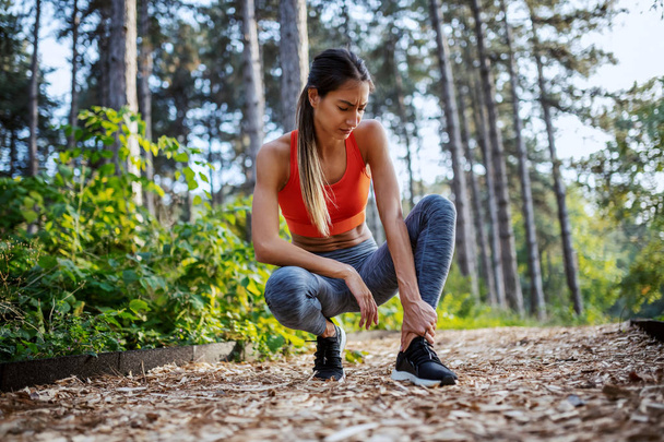 Fit attractive caucasian brunette in sportswear and with ponytail crouching on path in woods and holding injured ankle. Fitness in nature. - Photo, Image