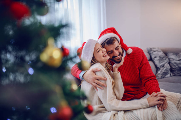 Encantador casal caucasiano em camisolas e com chapéu de Papai Noel na cabeça sentado no sofá na sala de estar ao lado da árvore de Natal e abraços
. - Foto, Imagem