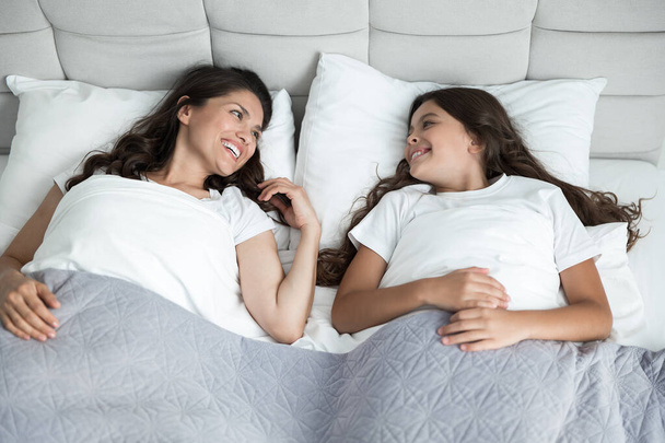 junge schöne Mutter und ihre süße Teenager-Tochter wachten gerade zusammen im Bett auf und lächelten einander zu - Foto, Bild