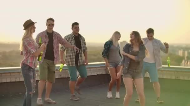 Egy hat fiatalból álló cég bulit rendezett a tetőn sörrel. Három fiatal férfi és két fiatal lány táncol kockás ingben. Fiatal szőke táncok az előtérben farmernadrágban - Felvétel, videó