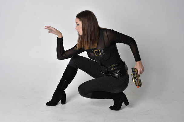 full length portrait of a pretty brunette woman wearing black leather fantasy costume. Sitting down with  a studio background. - Фото, зображення