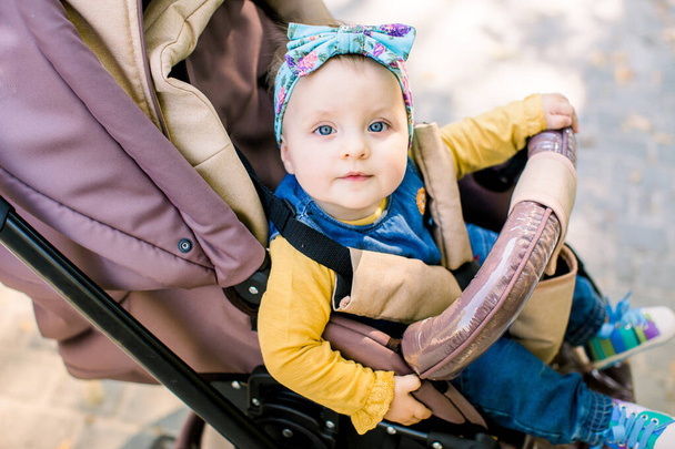 Happy girl in baby carriage playing in pram on background nature. Portrait cute little beautiful girl of 9 months sitting or stroller and waiting for mom. - Fotó, kép