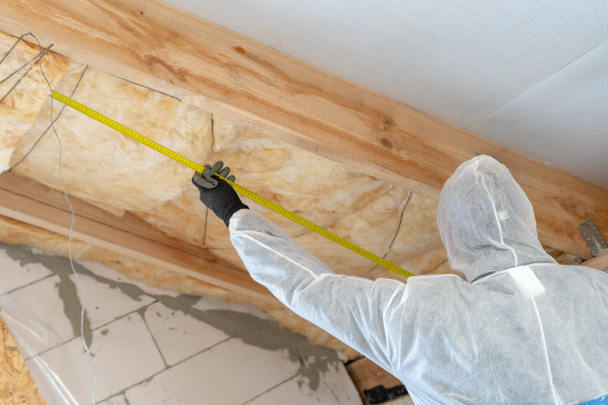 Constructor profesional hombre instalando capa de aislamiento térmico con lana de fibra de vidrio bajo el techo
 - Foto, Imagen