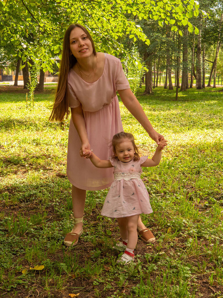 Mom and daughter walk and play in the park. Dressed in dresses. Sunny day, weekend in a city park. - Photo, Image