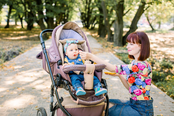 Mother Feeding Her Baby Girl with a Spoon. Mother Giving Food to her adorable Child . cute baby sitting on baby stroller carriage and posing smiling - Photo, Image