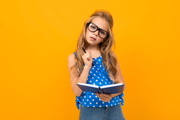 μικρό έξυπνο κορίτσι ποζάρουν με γυαλιά ηλίου και σημειωματάριο σε πορτοκαλί φόντο   - Φωτογραφία, εικόνα