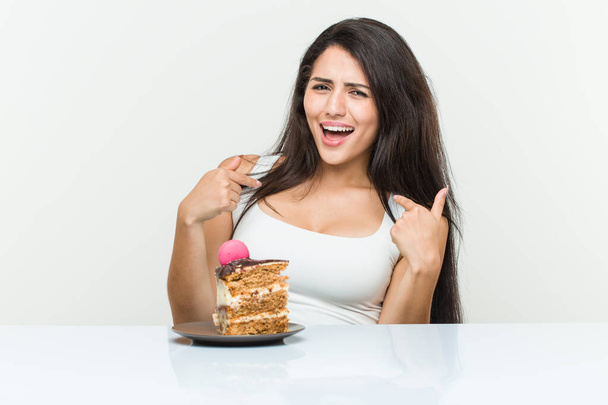 Young hispanic woman eating a cake surprised pointing at himself, smiling broadly. - Zdjęcie, obraz