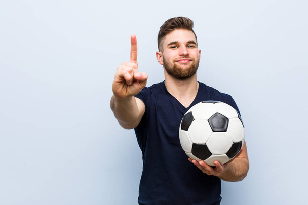 Jonge blanke man met een voetbal op nummer één met vinger. - Foto, afbeelding