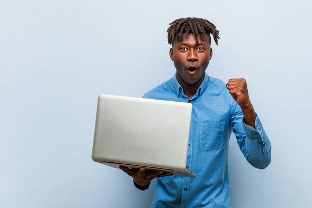 Young rasta black man holding a laptop cheering carefree and excited. Victory concept. - Foto, Imagem
