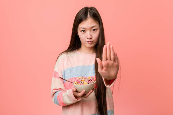 Νεαρή Ασιάτισσα με ένα μπολ δημητριακών στέκεται με απλωμένο χέρι που δείχνει στοπ, εμποδίζοντάς σας. - Φωτογραφία, εικόνα