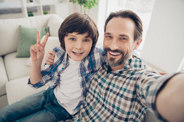 Comfortable weekend vacation with father concept. Close up photo of handsome bearded with teeth smile guy hugging his sweet son taking selfie making v-sign in living room house indoors - Photo, image
