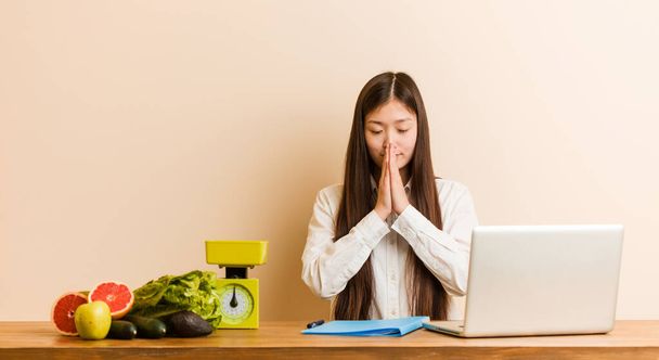 Joven nutricionista mujer china que trabaja con su portátil cogido de la mano en orar cerca de la boca, se siente seguro
. - Foto, imagen