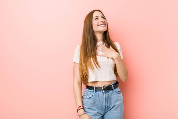 jonge roodharige vrouw tegen een roze muur lacht luid houden hand op de borst. - Foto, afbeelding