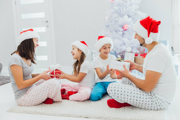 Family in santa s hats exchanging gifts in front of Christmas tree - Photo, image