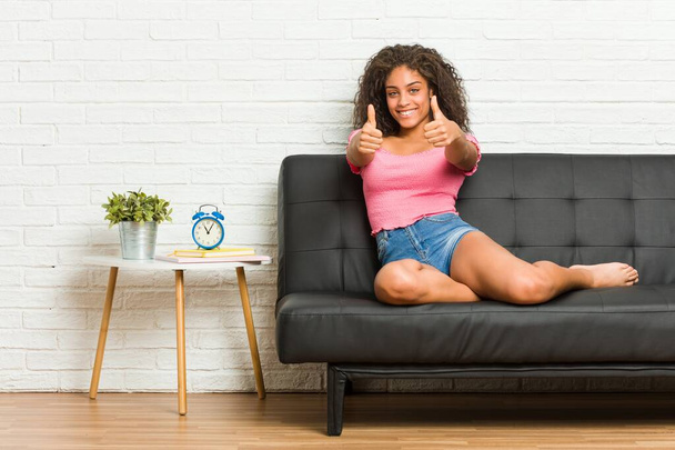 Młoda Afroamerykanka siedząca na kanapie z kciukami w górze, wiwatuje o czymś, wspiera i szanuje koncepcję. - Zdjęcie, obraz