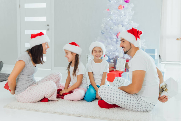 Family in Santa s hats exchanging gifts in front of Christmas tree - Foto, Imagen
