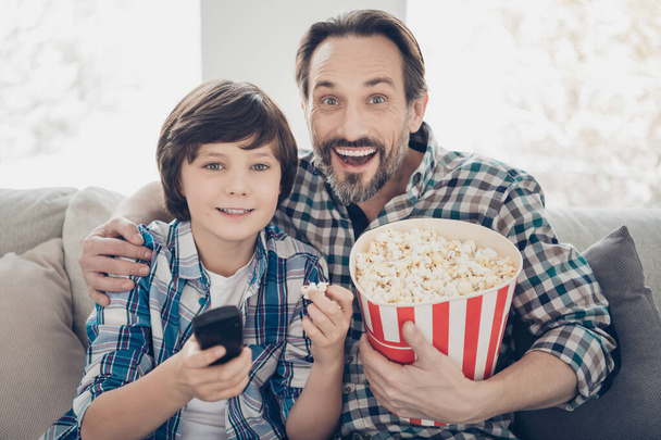 Close-up portrait of two nice attractive cheerful cheery guys sitting on sofa pre-teen son daddy hugging watching cartoon tv having fun eating snack in light white modern interior house living-room - Фото, зображення