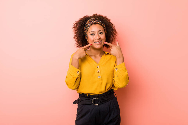 Νεαρή Αφροαμερικανή γυναίκα με ροζ φόντο χαμογελά, δείχνοντας με τα δάχτυλα στο στόμα. - Φωτογραφία, εικόνα