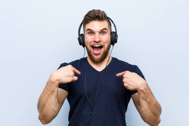 Jonge Kaukasische man die naar muziek luistert verrast wijzend naar zichzelf, breed lachend. - Foto, afbeelding