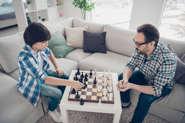 Portrait of two nice intelligent focused concentrated serious guys dad and pre-teen son sitting on sofa playing chess moving pieces in light white modern interior house living-room - Foto, immagini