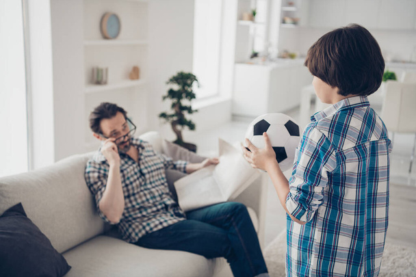 Pre-teen son asking inviting dad go play with him spend day. Father sitting on sofa reading finance press news in light white modern interior style studio house living-room - Фото, зображення